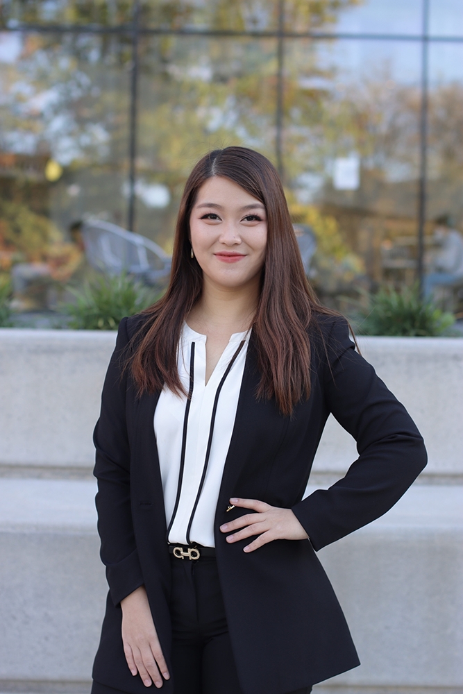 Photo of a smiling APIDA female student is a white blouse and black jacket smiling with a nature background.